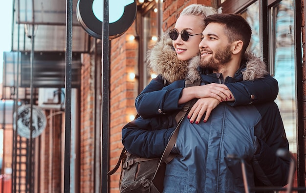 A happy attractive couple wearing winter coats is spending time together outdoors. A beautiful girl hugs her boyfriend while they are standing near the cafe outside.