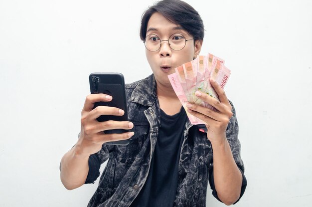 幸せな魅力的なインドネシアの若い男は、ルピアの紙幣と電話を持っている間、喜んでショックを受けました。