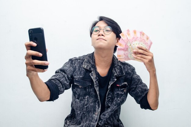 Happy attractive indonesian young man shocked happily while holding rupiah paper money and phone.