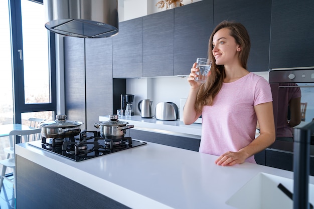 Happy attractive joyful brunette female drinking fresh clean filtered purified water at kitchen at home. Healthy lifestyle and quench thirst