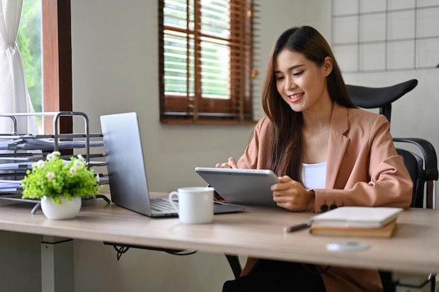 デジタル タブレットを使用して彼女の机で幸せで魅力的なミレニアル世代のアジアの実業家