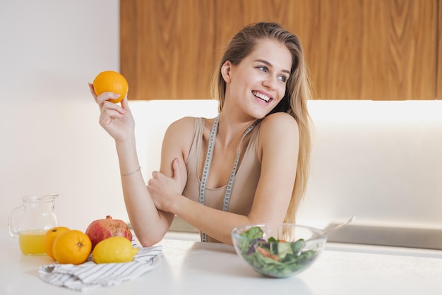 Happy attractive millennial woman in sportswear with measuring tape holding fresh orange for energy recovery after workout in kitchen