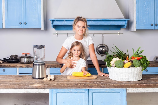 Happy attractive mother hugging daughter standing together in kitchen with modern furniture little girl holding corn basket of fresh vegetables on table vegan nutrition healthy food indoor