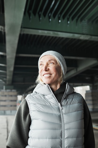 Happy attractive senior Caucasian woman in gray hat and vest looking aside and standing under city bridge outdoors