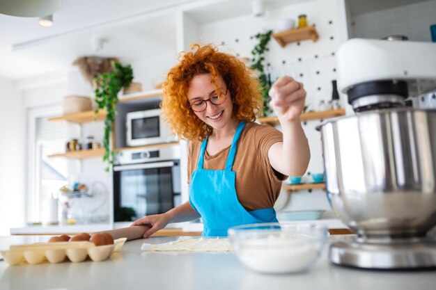 Happy attractive young adult woman housewife baker wear apron holding pin rolling dough on kitchen table baking pastry concept cooking cake biscuit doing bakery making homemade pizza at home
