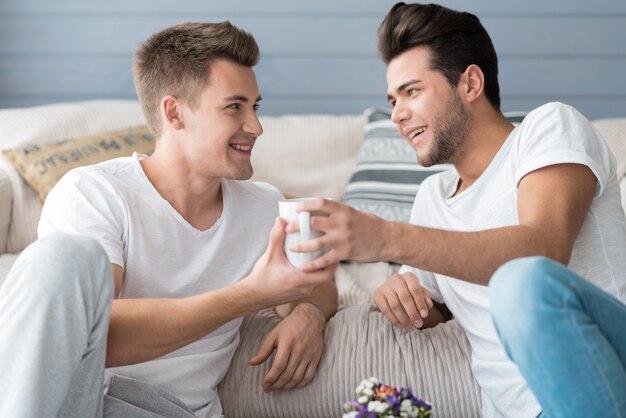 Happy attractive young gay couple smiling and drinking coffee while sitting on the floor