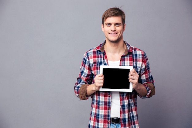 Happy attractive young man in plaid shirt standing and holding blank screen tablet over grey wall