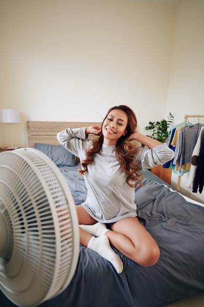 Happy attractive young vietnamese woman sitting on bed in front\
of blowing fan