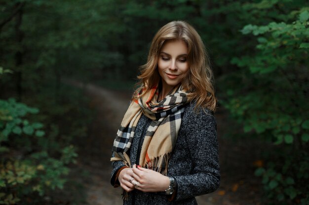 Happy attractive young woman in a fashionable gray coat with a stylish checkered scarf is standing on a path in the forest near green trees