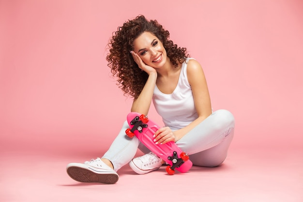 Happy attractive young woman sitting on skateboard over pink.