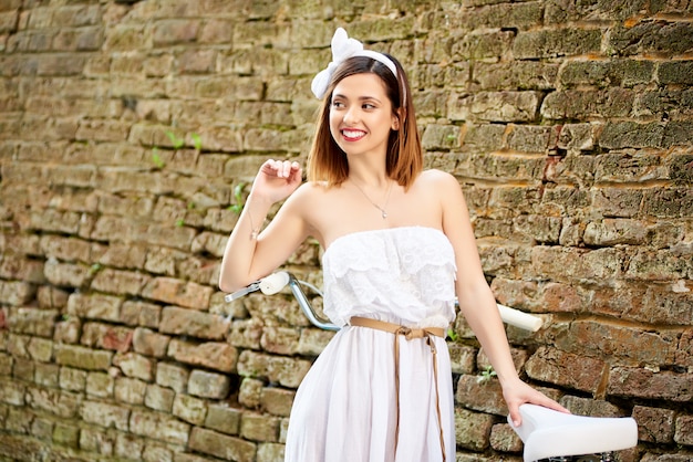 Happy attractive young woman smiling looking away joyfully standing near the brick wall in the city holding her bicycle