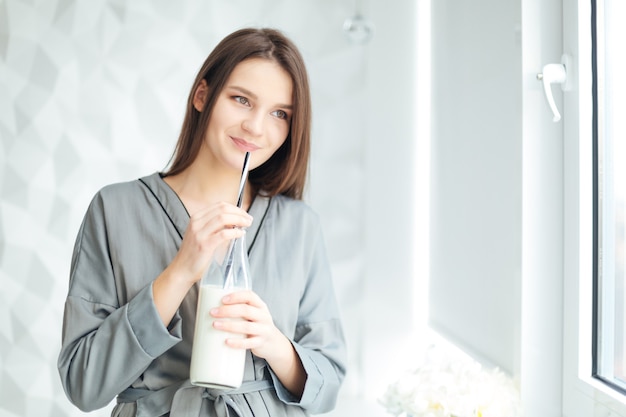 Happy attractive young woman standing near window and drinking milk