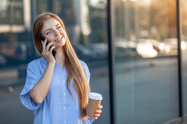 La giovane donna attraente felice che parla sullo smartphone, tenente porta via la tazza di caffè