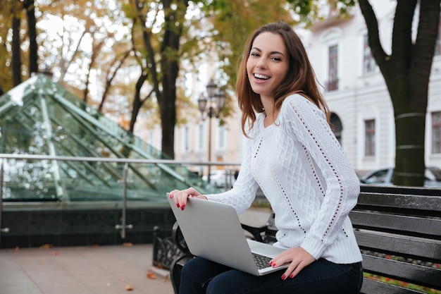 Happy attractive young woman using laptop on bench in park