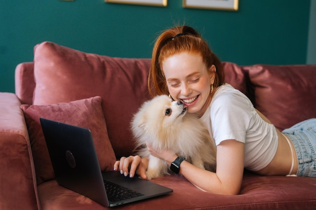 Happy attractvie young woman lying on comfortable sofa with white pretty Spitz pet dog and using laptop, closed eyes. Cute puppy licking nose of charming lady watching video from computer at home.