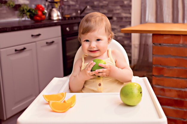 Happy baby 10-12 months eats fruit: orange, apple. Portrait of a happy girl in a highchair in the kitchen