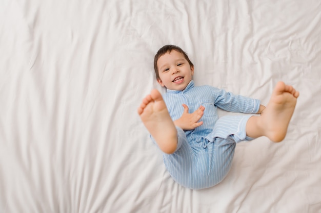 Happy baby boy in blue pajamas lies on the bed of the house, view from above, space for text