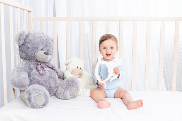 Happy baby boy in a crib in the nursery with a wooden toy laughing or smiling on a cotton white bed