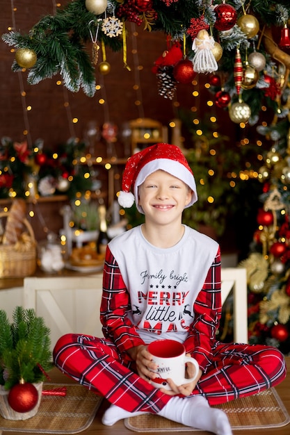 幸せな男の子が赤いクリスマスの装飾が施された台所のテーブルに座って、お茶を飲み、休日を見越して微笑んでいる