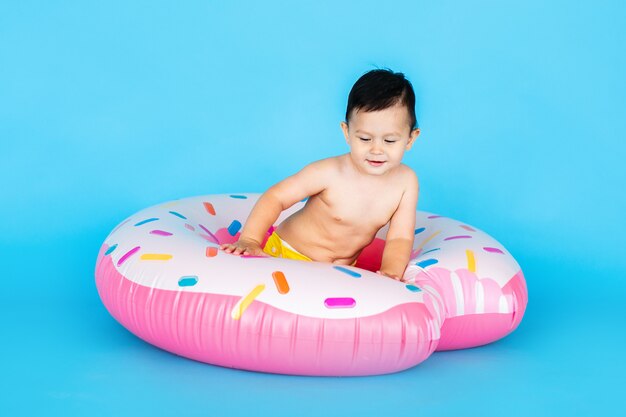 Happy baby boy in swimsuit with swimming ring donut on a colored blue wall