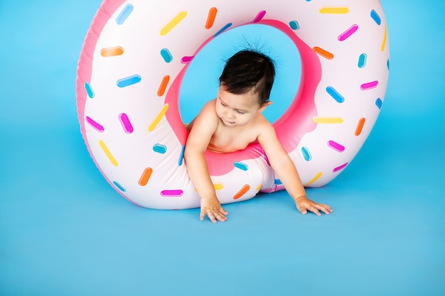 Happy baby boy in swimsuit with swimming ring donut on a colored blue wall