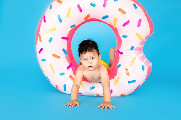 Happy baby boy in swimsuit with swimming ring donut on a colored blue wall