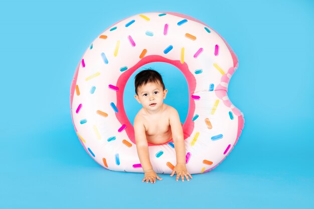 Happy baby boy in swimsuit with swimming ring donut on a colored blue wall