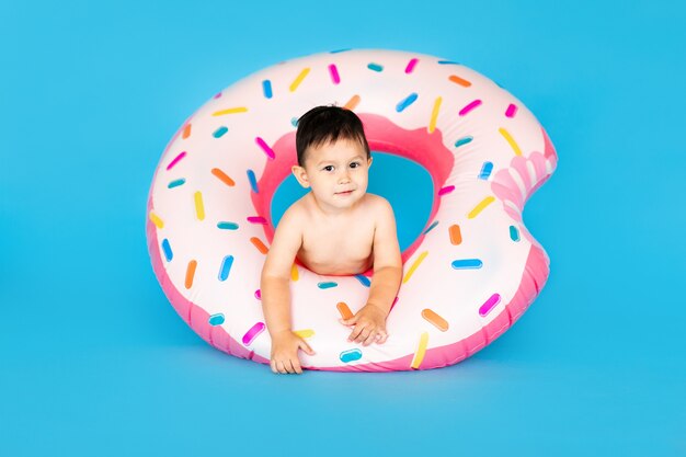 Happy baby boy in swimsuit with swimming ring donut on a colored blue wall