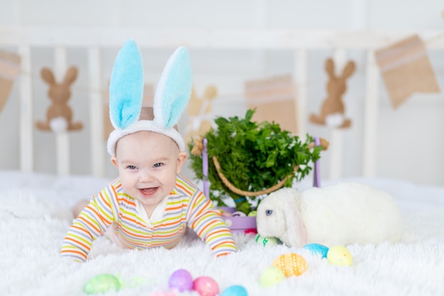 Happy baby boy with rabbit ears on his head lying with a rabbit on the bed with Easter eggs, cute funny smiling little baby. The concept of Easter.