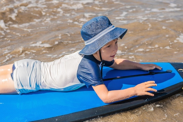 Photo happy baby boy - young surfer ride on surfboard with fun on sea waves. active family lifestyle, kids outdoor water sport lessons and swimming activity in surf camp. summer vacation with child