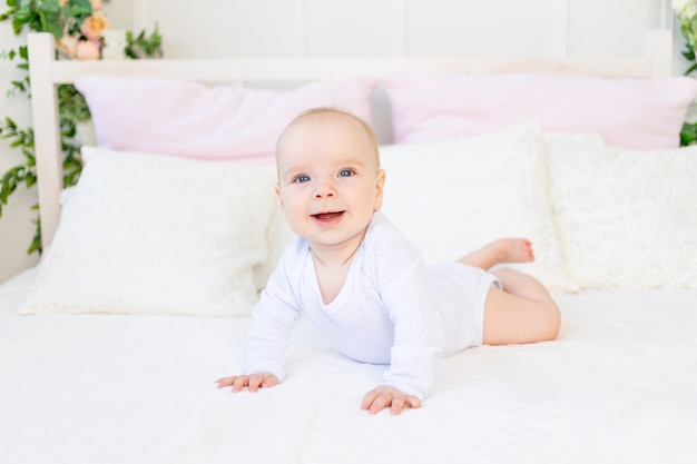 Happy baby girl 6 months old in a white bodysuit lying on her stomach on a white bed at home