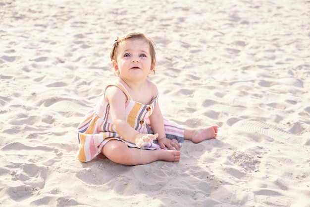 화창한 날 줄무늬 여름 드레스를 입고 모래 해변에 앉아 행복한 아기 소녀