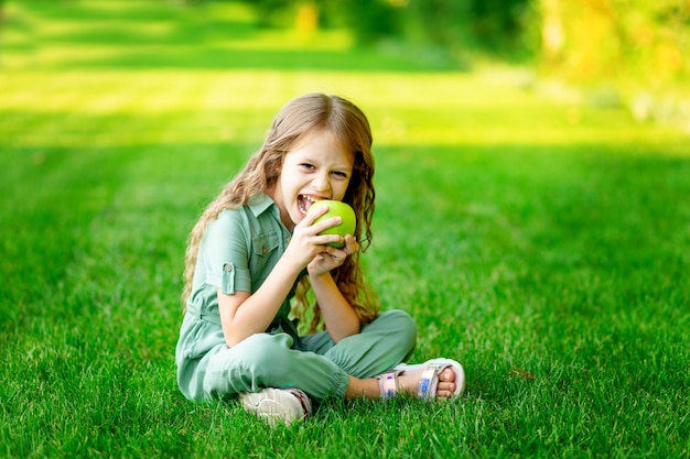 Una bambina felice in estate sul prato morde una mela verde con denti sani sull'erba e sorride, spazio per il testo