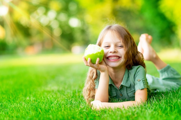 Happy baby girl in summer on the lawn bites a green apple with healthy teeth on the grass and smiles space for text