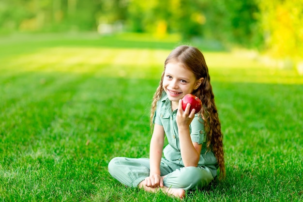 Happy baby girl in summer on the lawn with a red apple on the green grass and smiling space for text