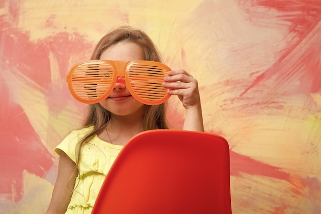안경을 입은 행복한 아기 소녀가 다채로운 배경에 의자에 앉아 있습니다.