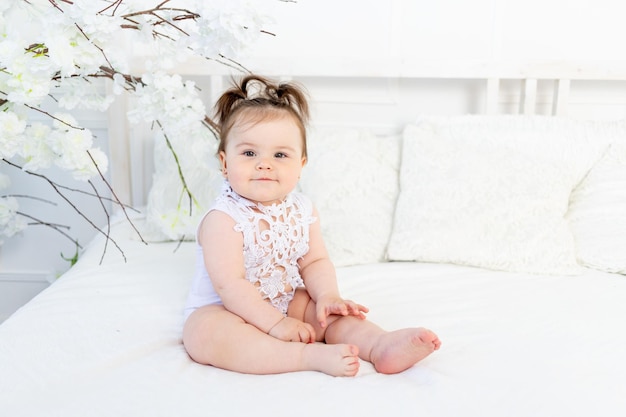 Happy baby girl in a white bodysuit on the bed at home in a bright room smiling