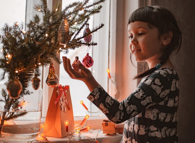 彼女の手に贈り物を持って笑っているクリスマスの窓で幸せな女の赤ちゃん。新年の願い事をしてください。