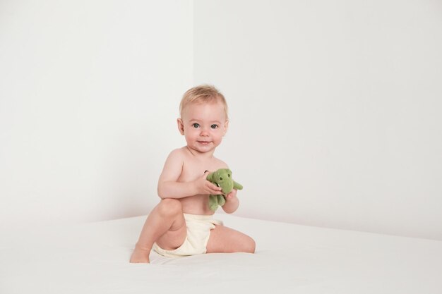 Happy baby is playing with green plush toy on white bed in white room