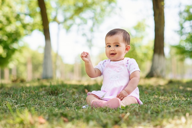 Happy baby in a pink dress sits on the grass in the park. Little baby girl plaing on the green grass.