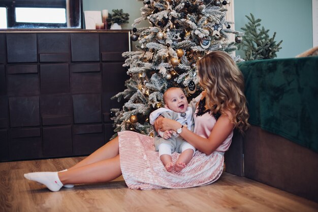 自宅で飾られたクリスマスツリーの横にある母親の腕の中で座っている幸せな赤ちゃん。