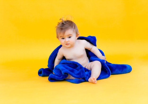 노란색 배경에 앉아 있는 행복한 아기는 후드가 있는 파란색 수건에 싸여 있습니다. 목욕 후 아기.