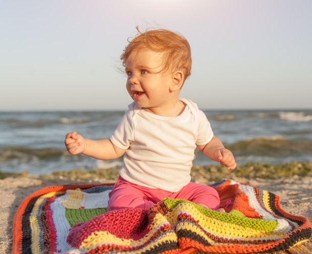 Happy baby smiling and waving hand, sitting on white sandy tropical beach on the carpet