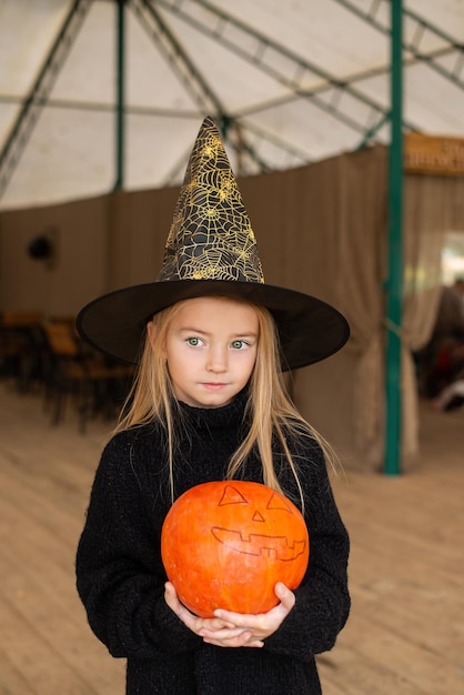 Happy baby in a witch hat for Halloween, pumpkins on the farm and hay