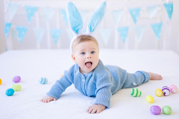 A happy baby with Easter bunny ears colorful Easter eggs lies at home on a bed on white pastel linen Easter Greeting Card
