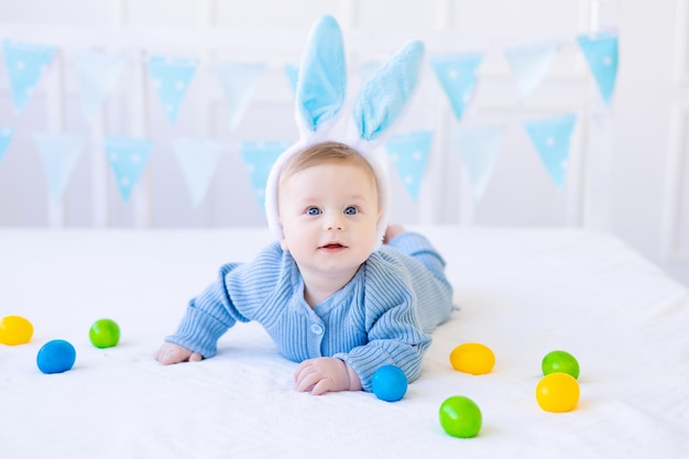 A happy baby with Easter bunny ears colorful Easter eggs lies at home on a bed on white pastel linen Easter Greeting Card