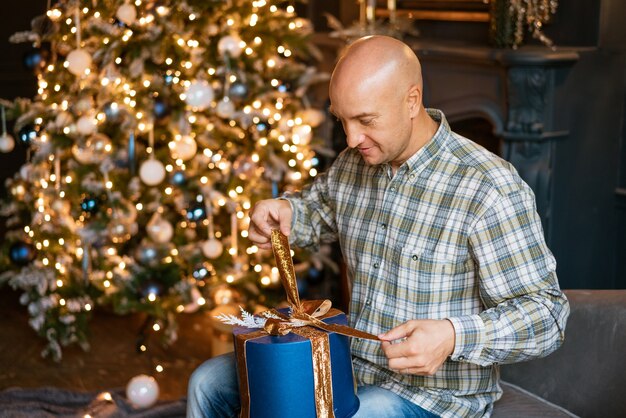 Felice uomo calvo in camicia apre un regalo di natale mentre è seduto sul divano sullo sfondo...