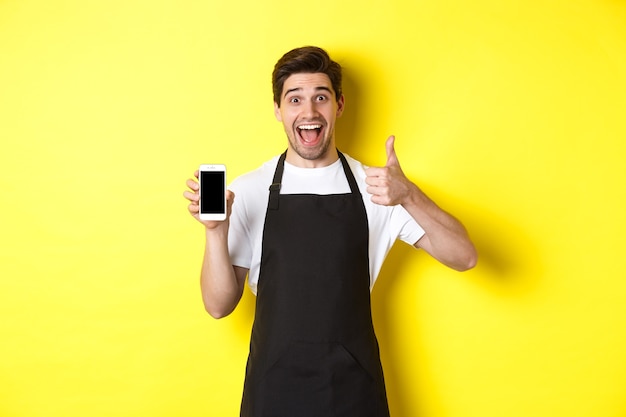 Happy barista in black apron showing smartphone screen, make thumb-up, recommending cafe application, standing over yellow wall