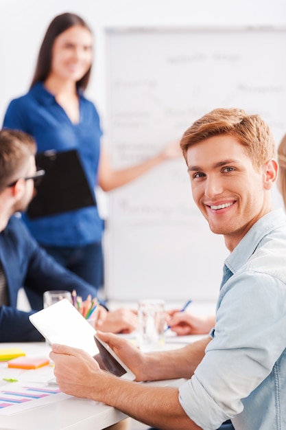 Happy to be a part of great team. Confident young man holding digital tablet and smiling while woman standing in the background and pointing whiteboard