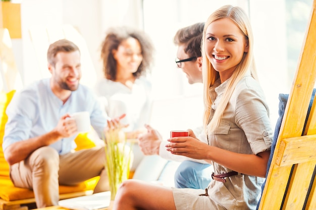 Felice di far parte di un team di successo. felice giovane donna che tiene una tazza di caffè e guarda la telecamera mentre i suoi colleghi discutono di qualcosa sullo sfondo